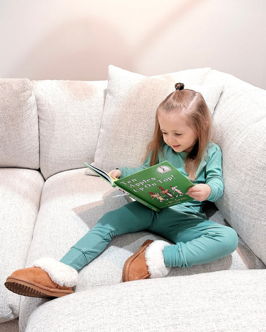 A young child with light brown hair, wearing the Dusty Teal Ribbed Solid • 2 Piece Bamboo Set (Updated Fit) thats gentle on sensitive skin, sits on a light sofa smiling while reading the green childrens book Ten Things To Spot.