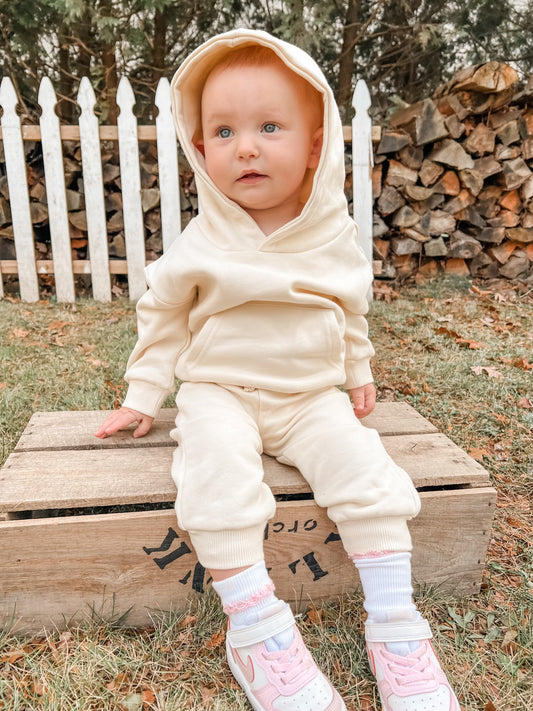 A baby in the Oatmeal • Sweatpant & Hoodie Set sits on a wooden crate outside, with a white picket fence and stacked firewood behind. The baby has blue eyes, light hair, and wears pink and white sneakers.