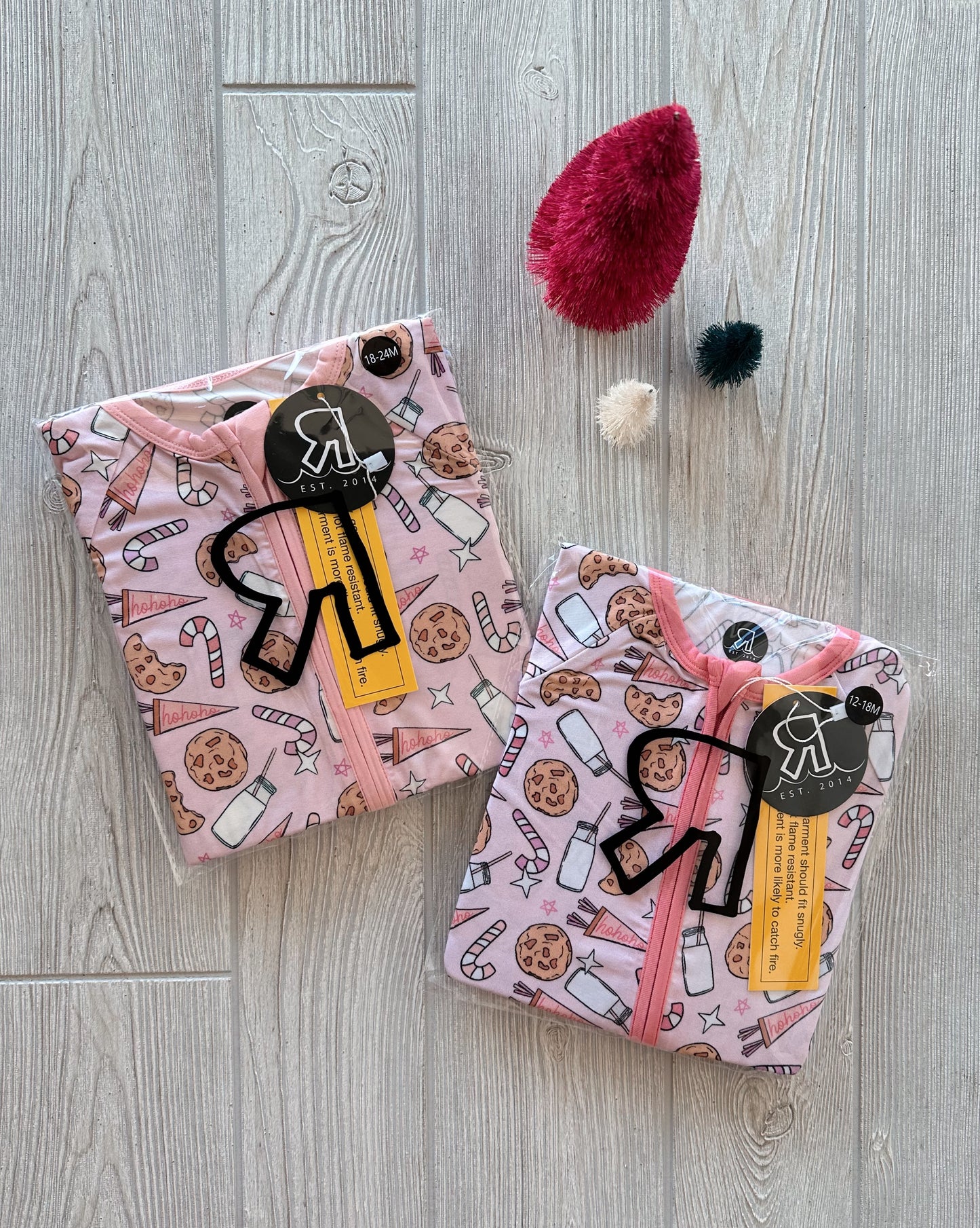 Two folded Milk & Cookies • Bamboo Zippy pajamas in light pink and lavender, featuring milk, cookie, and candy prints, rest on a light wood floor beside three fuzzy red, white, and black decorative trees.