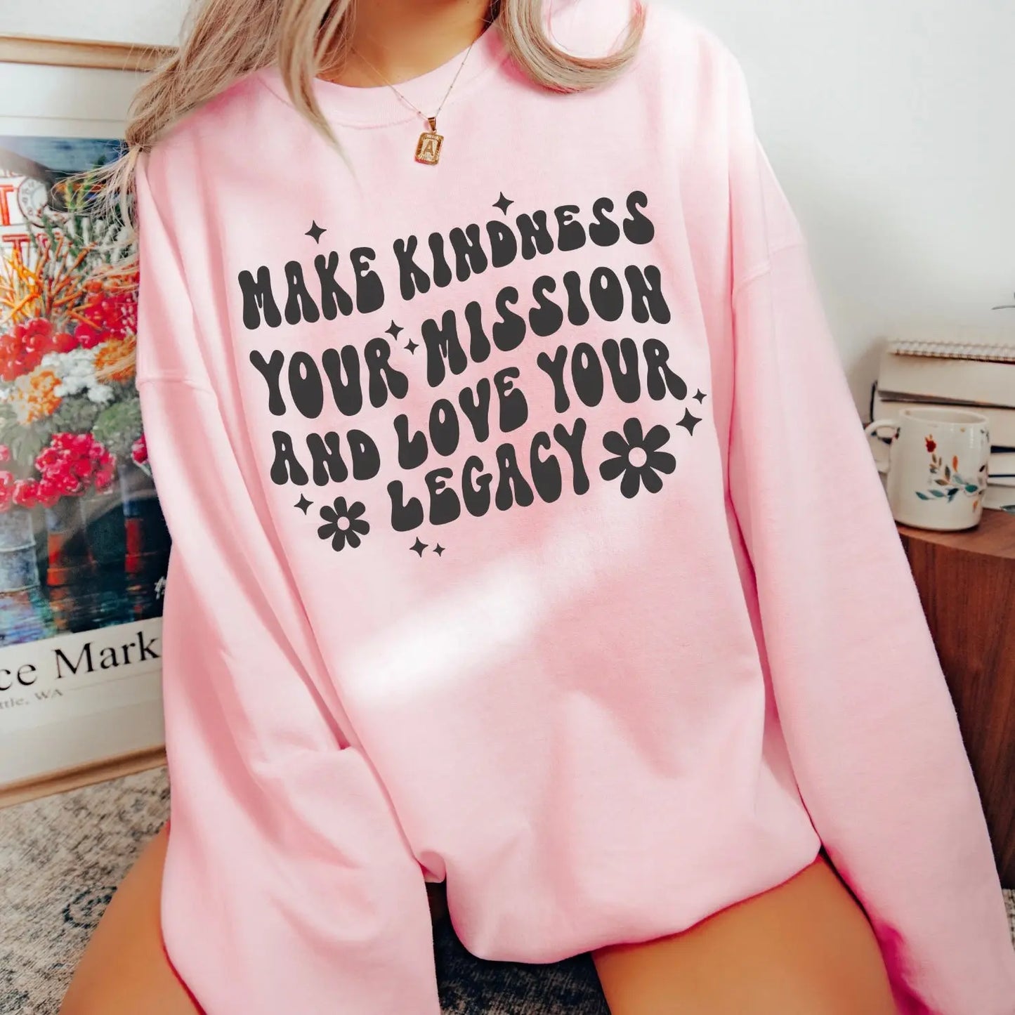 A person wears the Legacy • Baby Pink Pullover, a light pink unisex sweatshirt featuring bold black text, “Make kindness your mission and love your legacy,” with small flowers and sparkles. Soft cotton/polyester blend.