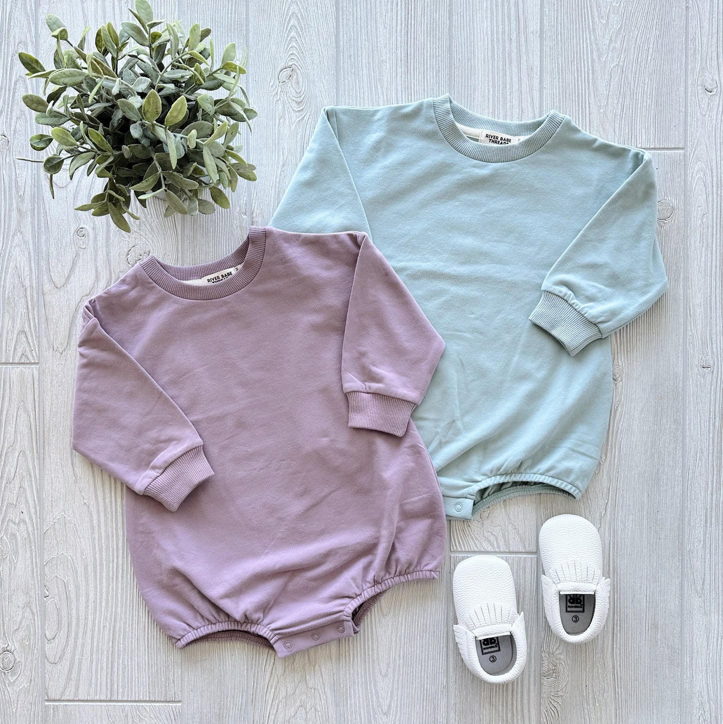 Two Spring Bubble Rompers made from organic cotton French terry—one lavender, one light blue—are displayed on a light wood floor. A small potted plant is on the left, with white baby shoes beside the blue romper. Perfect for cool weather.