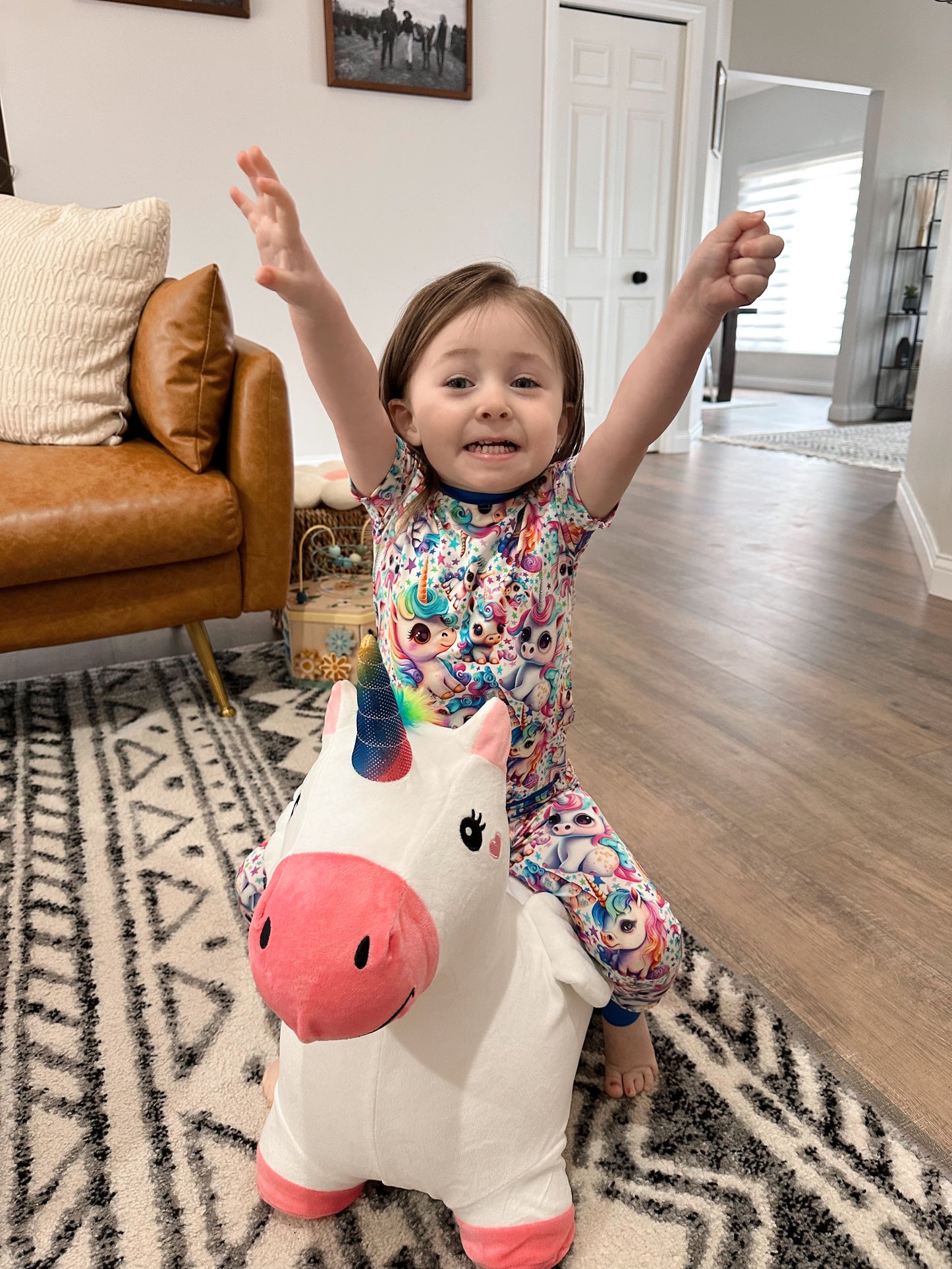 A young child in the Mythical Unicorns • 2 Piece Bamboo Set (Updated Fit) smiles with arms raised, sitting on a plush unicorn toy—colorful, comfy pajamas perfect for sensitive skin.