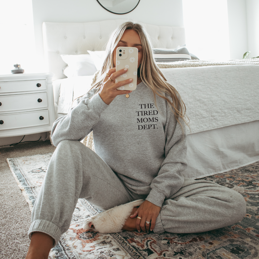 A woman sits on a rug in a cozy bedroom, wearing The Tired Moms Dept. Gray Pullover as she holds her phone up, the classic unisex sweatshirt featuring soft cotton/polyester fabric and bold lettering across the front.