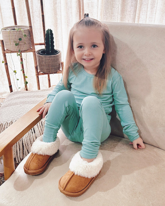 A young girl with long blonde hair, wearing the Dusty Teal Ribbed Solid • 2 Piece Bamboo Set (Updated Fit) and oversized brown slippers with white fur, sits smiling on a beige armchair with houseplants and a wooden screen in the background.