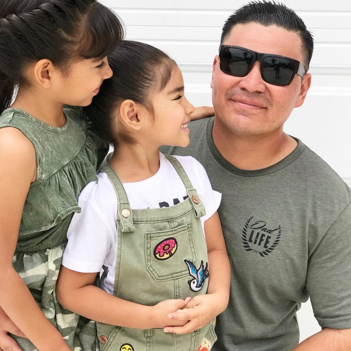 A man in sunglasses and a Dad Crest • Heather Olive Tee smiles while posing with two young girls in patterned green outfits, laughing and facing each other, all against a white background.