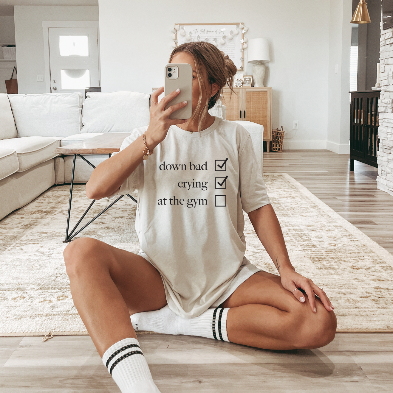 A woman sits cross-legged on a rug, taking a mirror selfie in her cozy living room. She wears the Not at the Gym cream tee—ideal for fans of comfy, retail fit style with a playful touch.