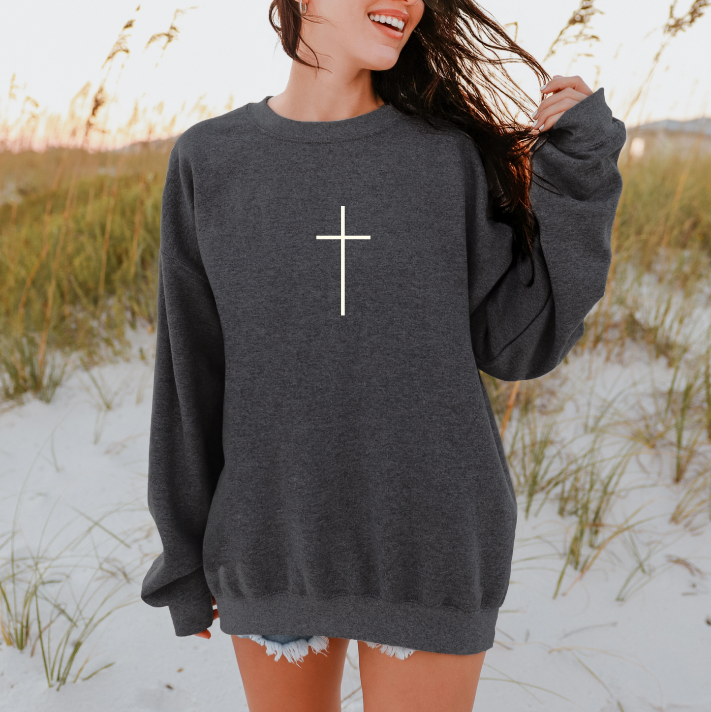 A woman smiles and touches her hair on a sandy beach, wearing the Faith Over Fear Charcoal Tee with a white cross design. The unisex tee fits true to size; tall grass and blue water are in the background.