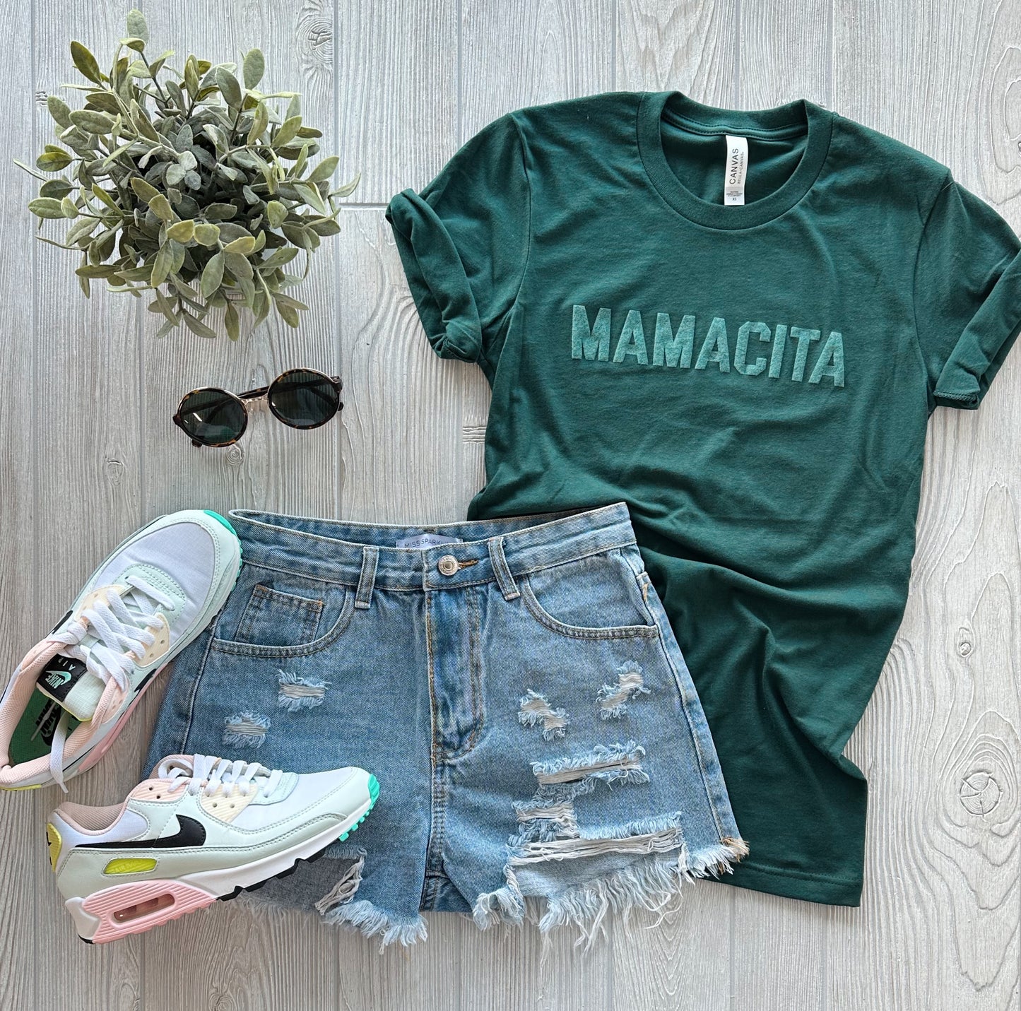 A flat lay of a MAMACITA • Forest Tonal Bella Canvas comfy mom tee, distressed denim shorts, white pastel-accented sneakers, round sunglasses, and a small potted plant on a light wood background.
