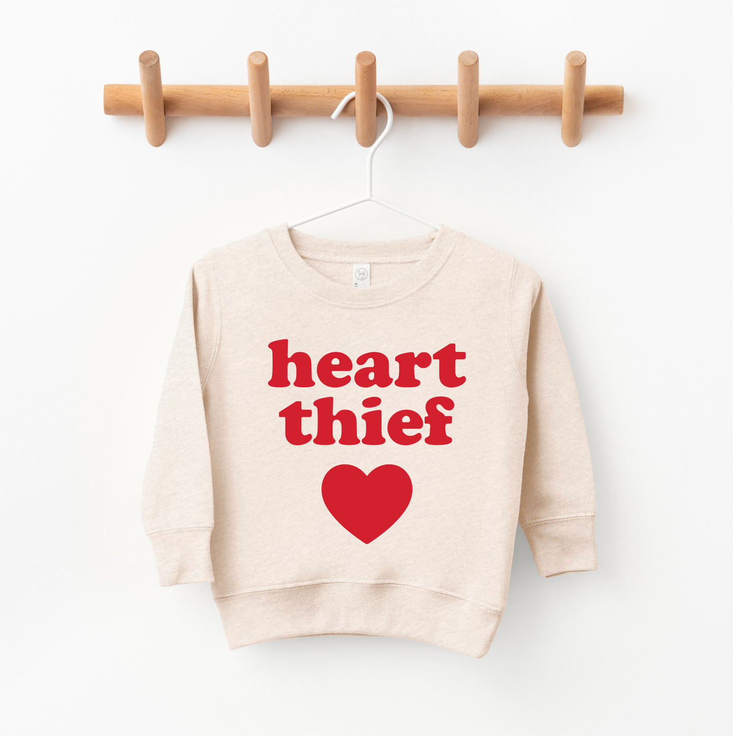 The Heart Thief • Oatmeal Pullover is a beige unisex sweatshirt with bold red heart thief text above a red heart graphic. Shown on a white hanger against a wooden wall rack. For extra coziness, we recommend sizing up.