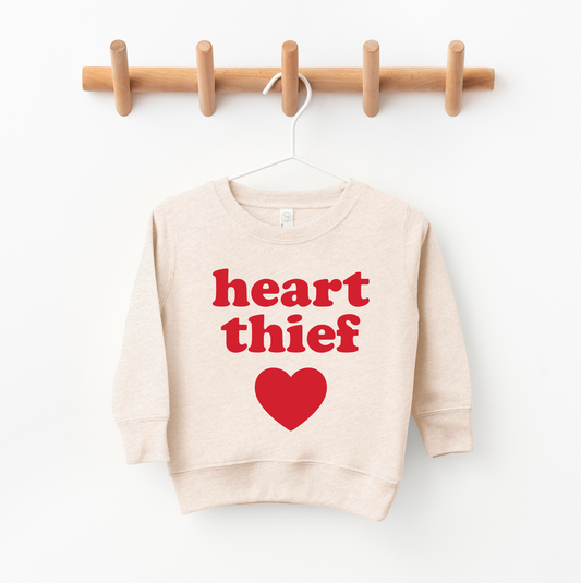 The Heart Thief • Oatmeal Pullover is a beige unisex sweatshirt with bold red heart thief text above a red heart graphic. Shown on a white hanger against a wooden wall rack. For extra coziness, we recommend sizing up.