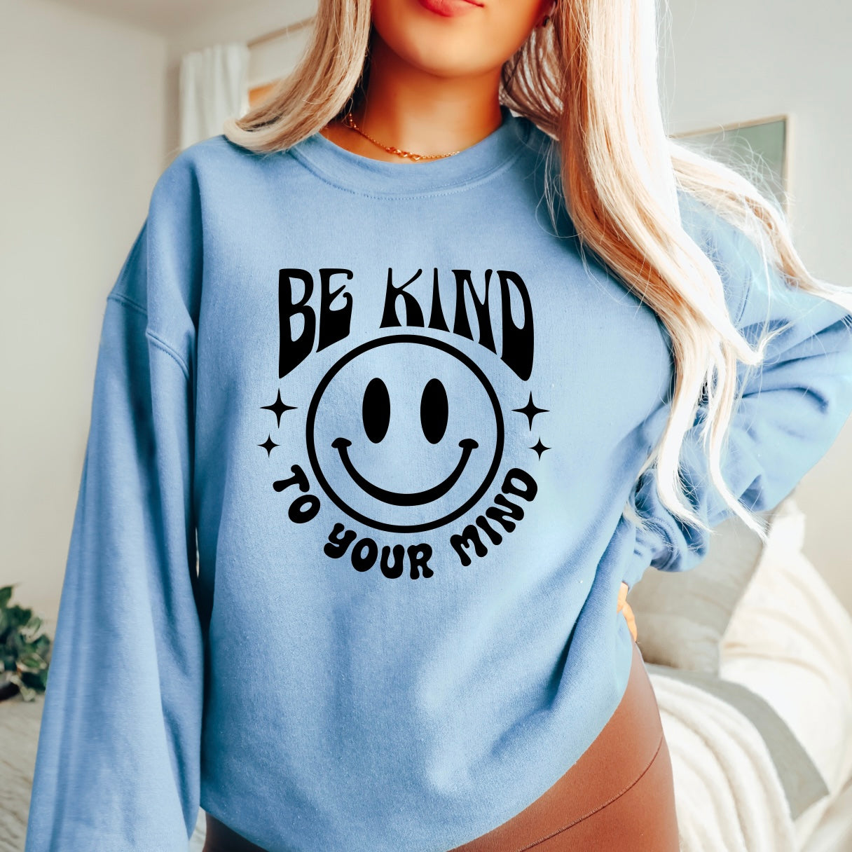 Someone stands in a cozy, well-lit room wearing the Kind to Your Mind • Blue Pullover, a classic fit unisex sweatshirt featuring Be Kind To Your Mind and a large smiley face, made from soft cotton polyester.