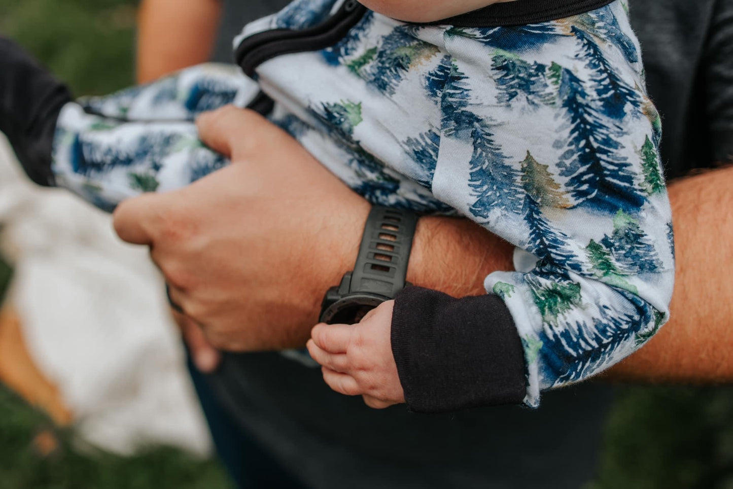 A close-up shows an adult cradling a baby in the Snowy Trees • Bamboo Zippy (Updated Fit) with a blue and green pine tree pattern, creating a warm, cozy scene. The adult wears a dark watch; the background is softly blurred for added warmth.