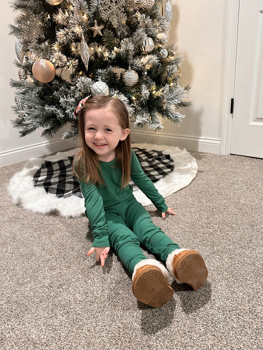 A young girl sits smiling on the carpet in front of a snowy Christmas tree with white and gold ornaments. She wears a bow in her hair, fuzzy boots, and the Forest Green Ribbed Solid • 2 Piece Bamboo Set (Updated Fit).