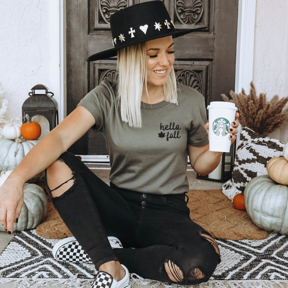 A woman in a black wide-brim hat and a fitted hello fall • Army Tee sits cross-legged on a patterned rug, holding a Starbucks cup, surrounded by pumpkins and cozy fall decor.