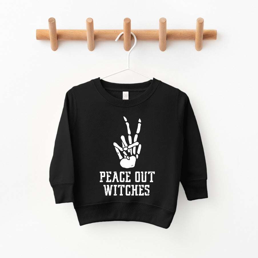 The Peace Out Witches • Kids Pullover is a black, oversized unisex sweatshirt featuring a white skeleton hand peace sign above PEACE OUT WITCHES in bold on the front, shown hanging on a wooden peg rack.