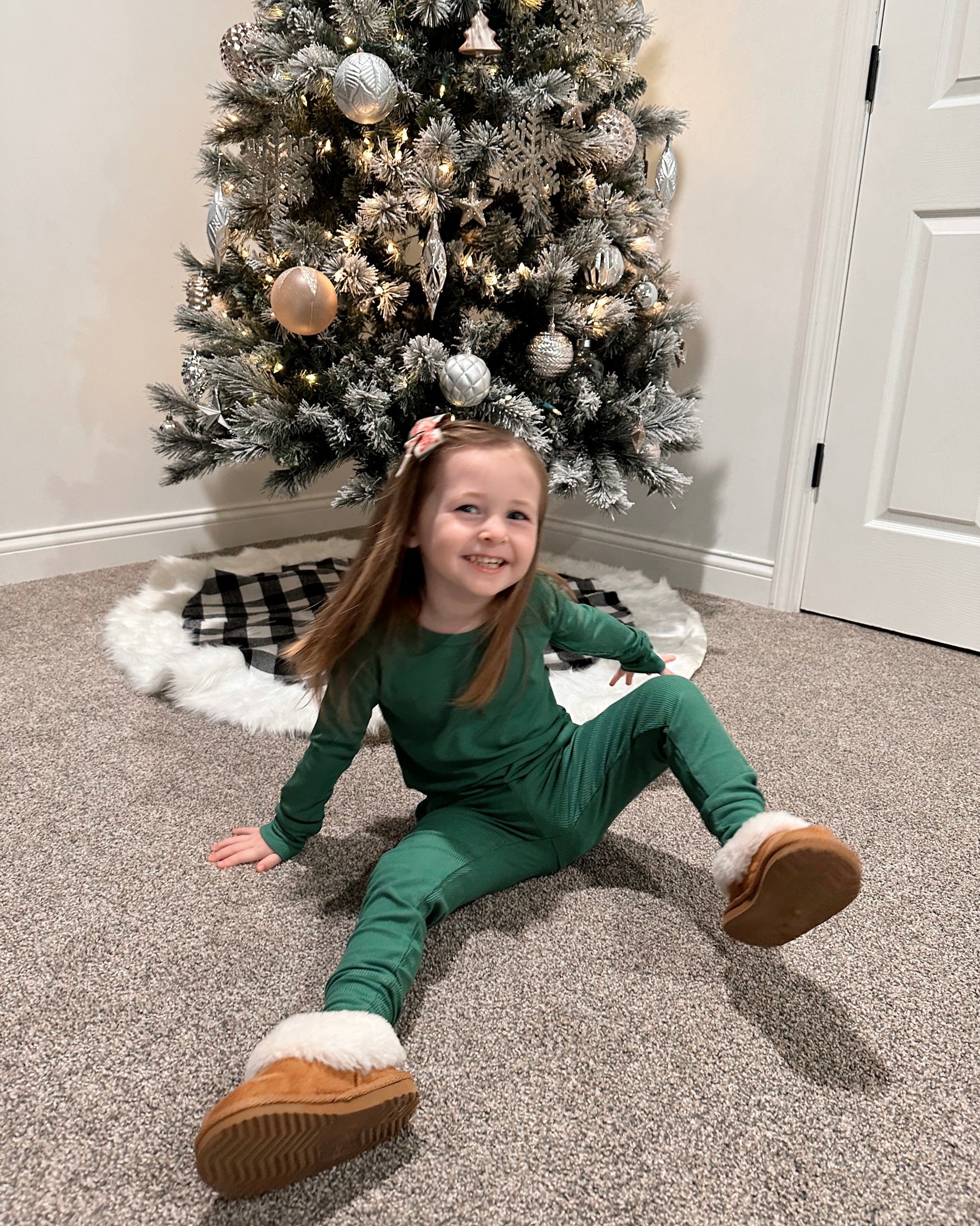 A young girl wearing the Forest Green Ribbed Solid 2 Piece Bamboo Set (Updated Fit) smiles while sitting on a carpet in front of a Christmas tree adorned with white and silver ornaments.