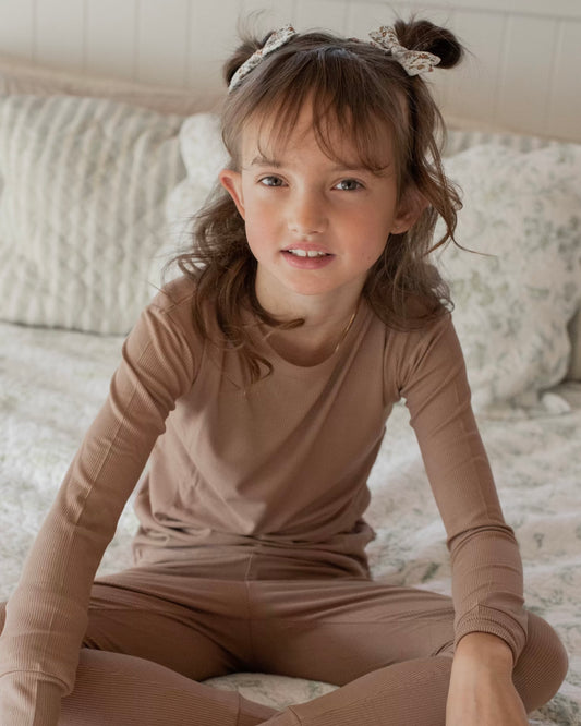 A young girl with brown hair in two buns and a bow headband sits cross-legged on a bed, smiling at the camera. She wears the Chestnut Ribbed Solid • 2 Piece Bamboo Set (Updated Fit), perfect for sensitive skin. The bedding is light and patterned.