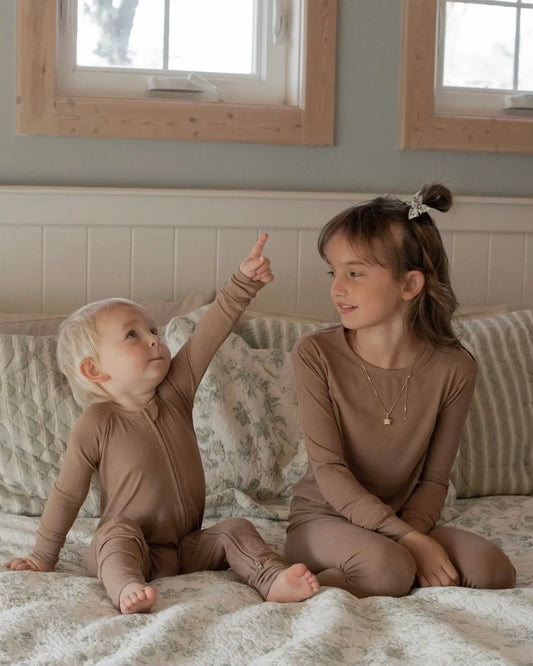 Two children sit on a bed in matching Chestnut Ribbed Solid • 2 Piece Bamboo Sets (Updated Fit), perfect for sensitive skin. The toddler points upward while the older child smiles, with soft bedding and sunlight streaming through the windows.