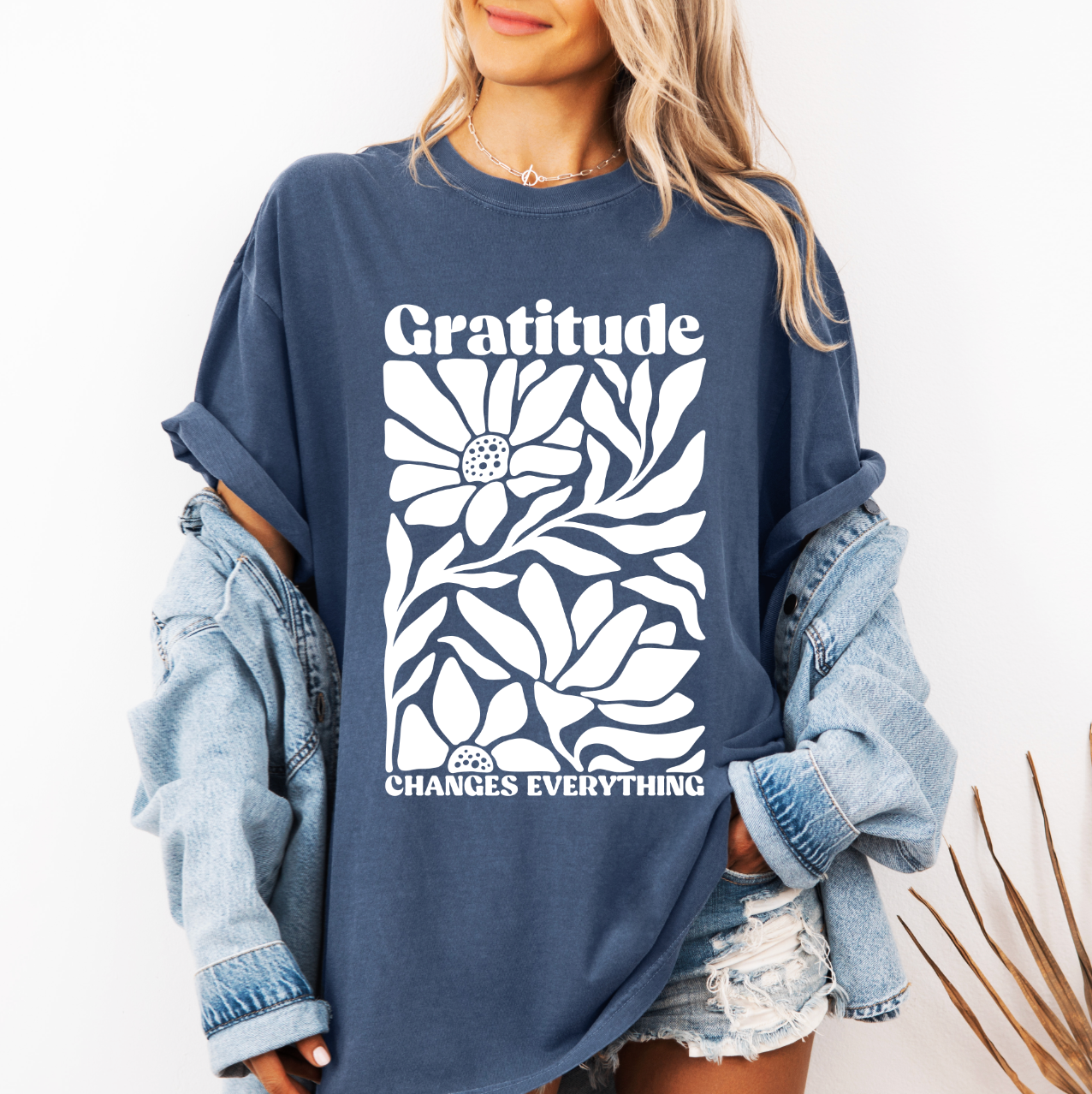 A woman wears the Gratitude • Comfort Colors Tee, a garment-dyed blue shirt with a floral graphic and Gratitude Changes Everything text, styled with a denim jacket and distressed denim shorts.