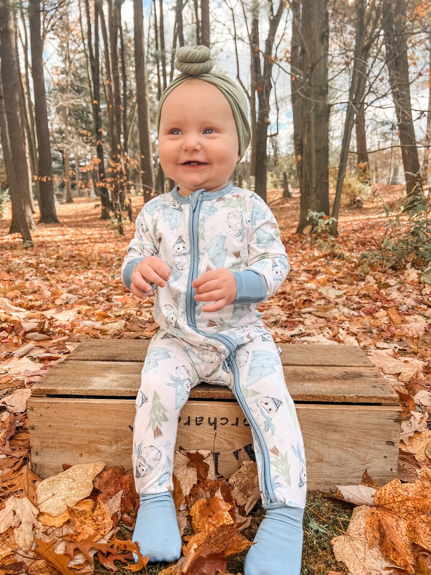 A smiling baby in the Abominable Snowman • Bamboo Zippy (Updated Fit) onesie and green headwrap sits on a wooden crate in a forest of autumn leaves, surrounded by tall trees and soft sunlight.