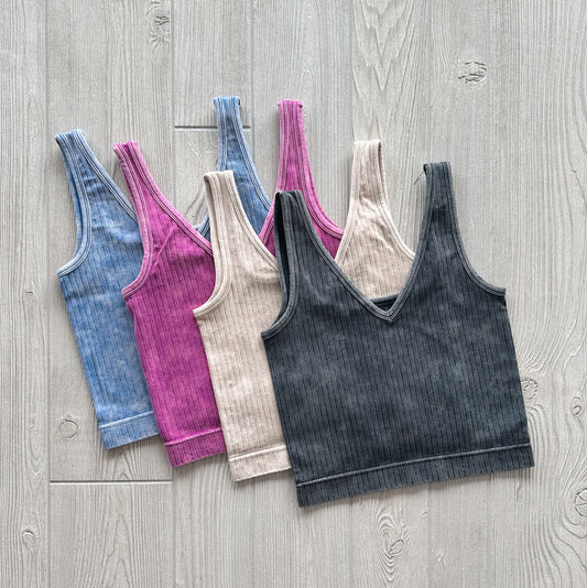 Four Ava Ribbed Crop bramis from the Vintage Collection—blue, pink, beige, and black—are fanned out on a light wooden surface, highlighting their soft, stretchy fabric.