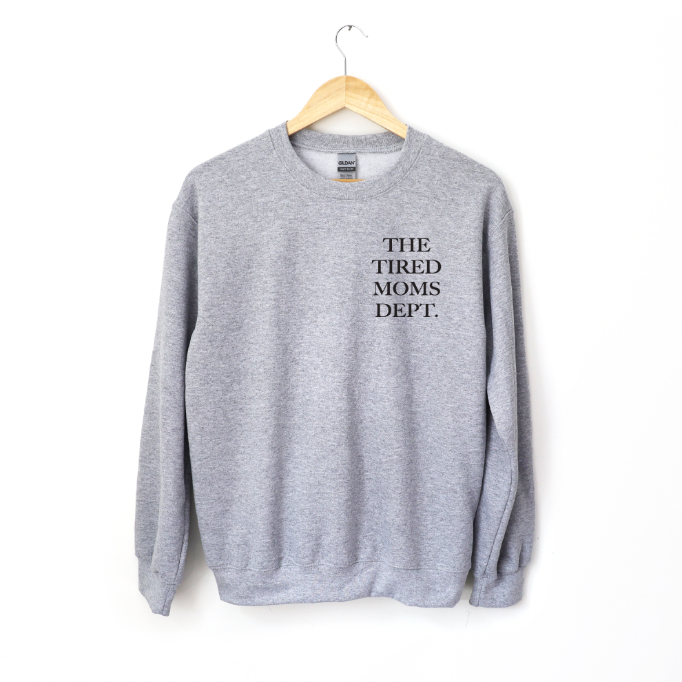 The Tired Moms Dept. • Gray Pullover is a classic fit crewneck sweatshirt in gray, featuring THE TIRED MOMS DEPT. in black on the upper left chest. Made from a cozy cotton/polyester blend and shown on a wooden hanger against a white background.