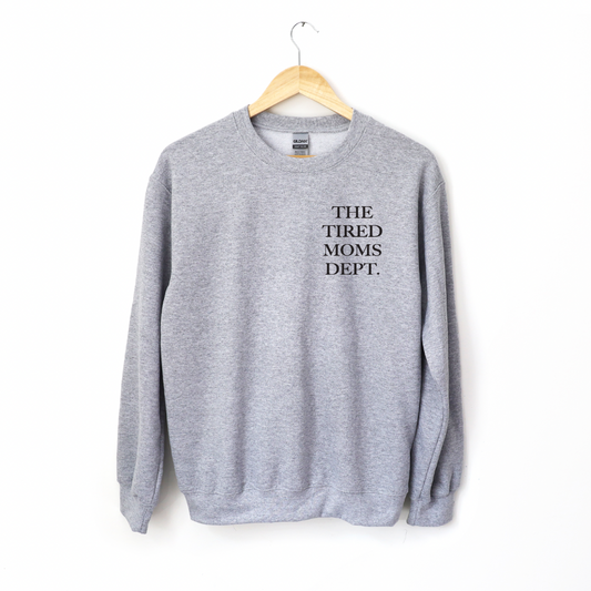 The Tired Moms Dept. • Gray Pullover is a classic fit crewneck sweatshirt in gray, featuring THE TIRED MOMS DEPT. in black on the upper left chest. Made from a cozy cotton/polyester blend and shown on a wooden hanger against a white background.