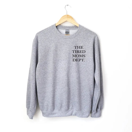The Tired Moms Dept. • Gray Pullover is a classic fit crewneck sweatshirt in gray, featuring THE TIRED MOMS DEPT. in black on the upper left chest. Made from a cozy cotton/polyester blend and shown on a wooden hanger against a white background.