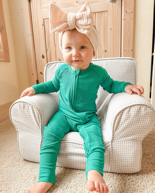 A baby wearing a large cream-colored bow headband and the Emerald Ribbed Solid • Bamboo Zippy (Updated Fit) sits on a small plaid armchair, looking at the camera in a cozy, light-filled room.
