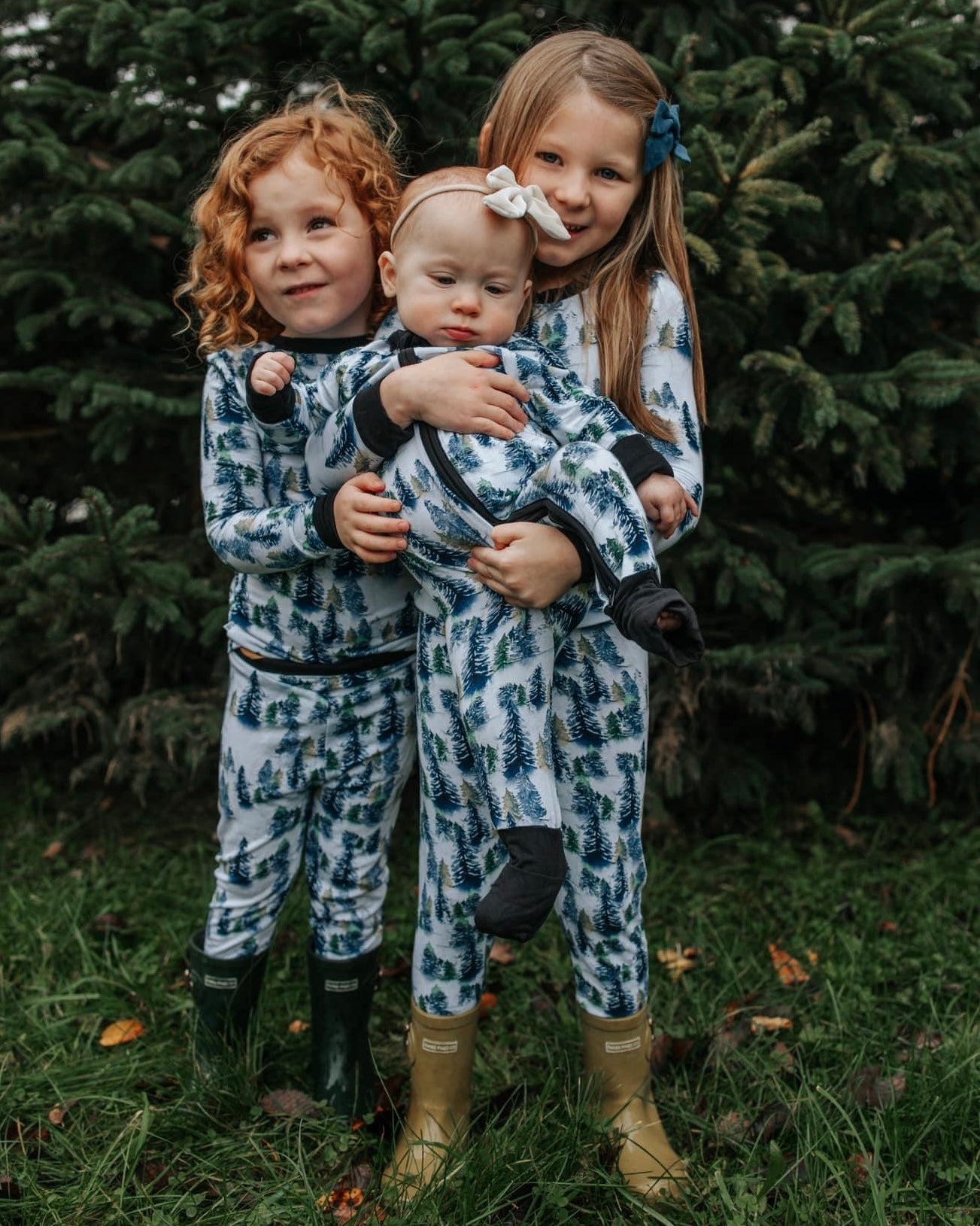 Three young children smile in front of pine trees, all wearing matching Snowy Trees • 2 Piece Bamboo Sets (Updated Fit) with a tree pattern. The oldest girl holds a baby, while the third stands by them on the grass; all are in rain boots.