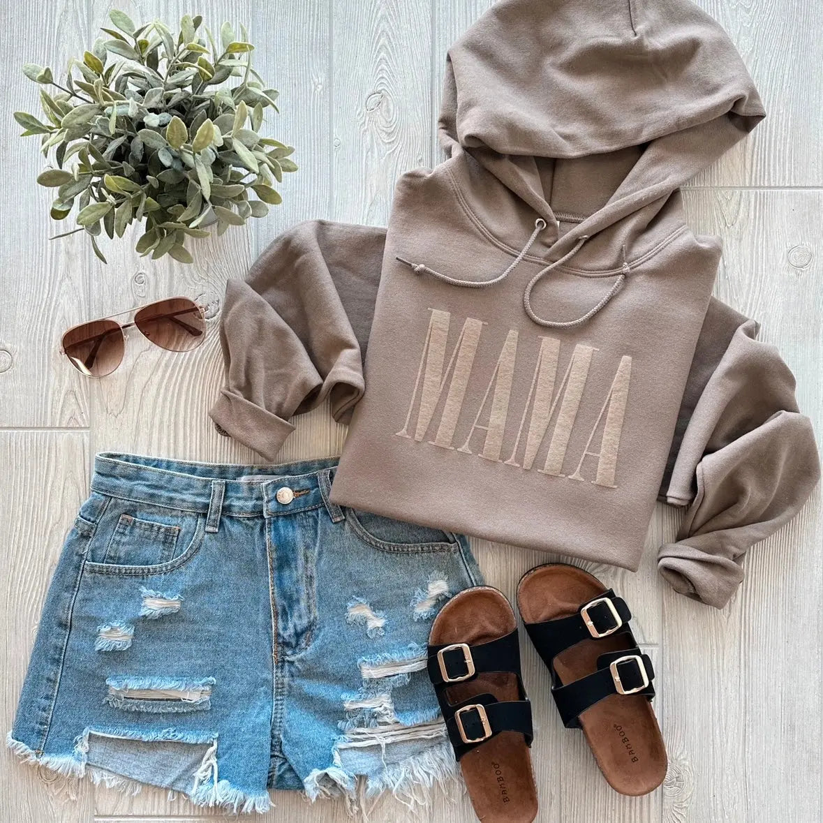 Flat lay featuring the MAMA • Latte Tonal Hoodie, paired with distressed denim shorts, black buckle sandals, brown sunglasses, and a small potted plant—all displayed on a light wood floor.