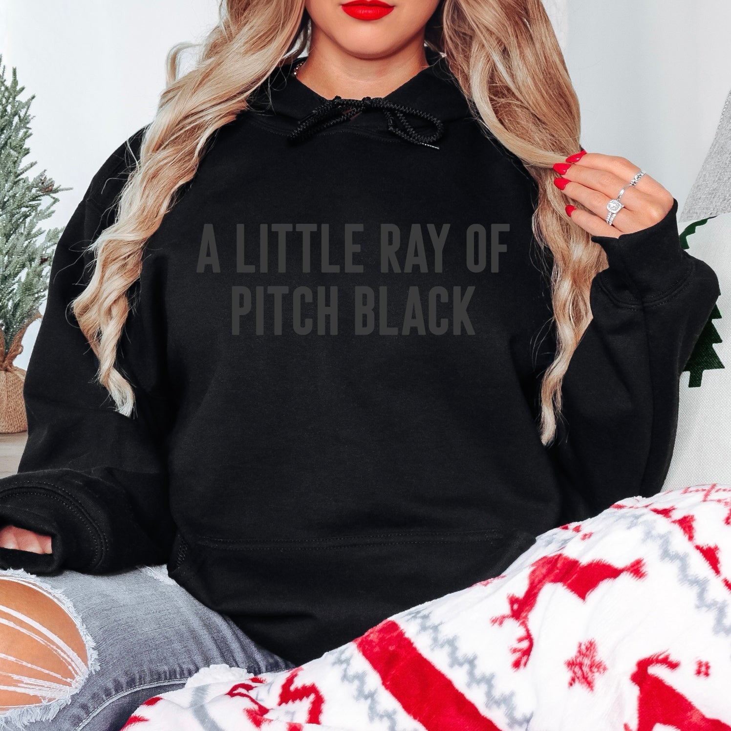 A woman with long blonde hair wears the Ray of Pitch Black • Tonal Hoodie, featuring A LITTLE RAY OF PITCH BLACK text. Sitting on a couch with a red and white reindeer blanket, she shows off the hoodie’s comfy unisex fit.