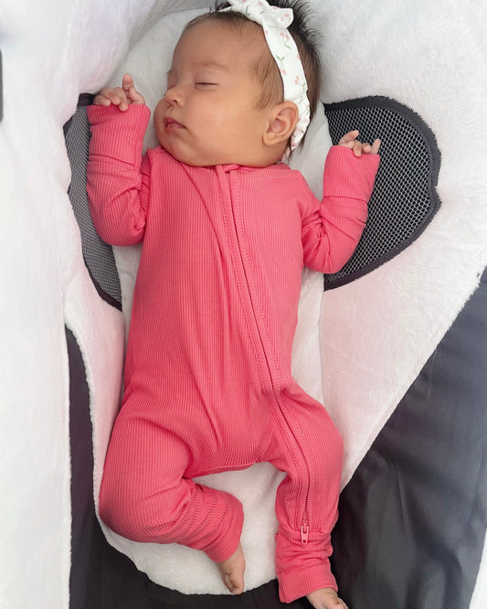 A baby sleeps peacefully on a soft, white and gray cushion, wearing the Bright Pink Ribbed Solid • Bamboo Zippy (Updated Fit), a gentle bamboo zip-up onesie, with a floral headband.