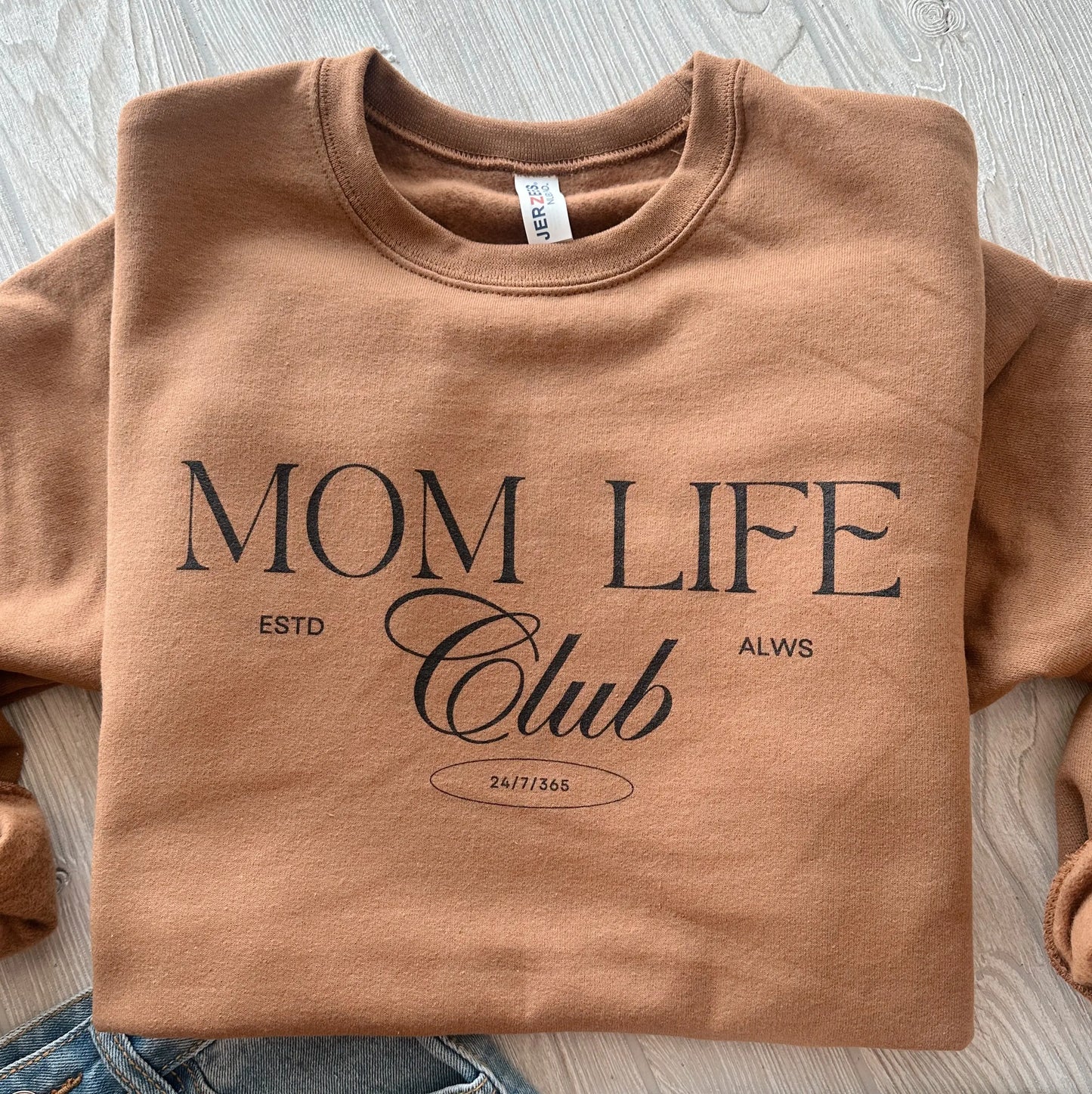The Mom Life Club • Caramel Pullover, a unisex-fit sweatshirt with "MOM LIFE Club ESTD ALWS 24/7/365" in black on the front, is folded on a light wooden surface beside denim jeans.