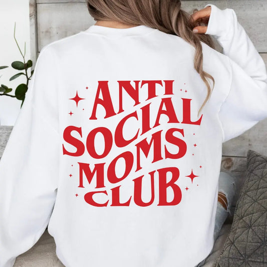 A woman sits indoors with her back to the camera, wearing the Anti-Social • Red & White Crew, a classic fit unisex fleece sweatshirt featuring bold ANTI SOCIAL MOMS CLUB text in red and white across the back.