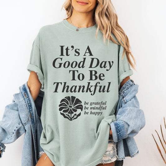 A woman wears a light green Good Day to be Thankful • Comfort Colors Tee made from ring-spun cotton with rolled sleeves, paired with a denim jacket. The shirt features “Its a Good Day To Be Thankful,” “be grateful be mindful be happy,” and a floral graphic.