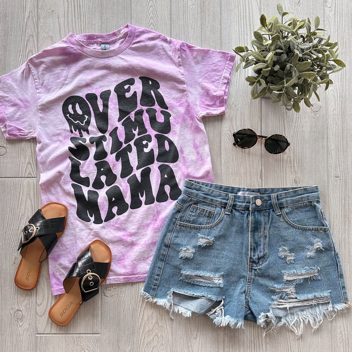 A flat lay of the Overstimulated • Lilac Tie Dye unisex tee with bold black text, paired with distressed denim shorts, black sandals, round sunglasses, and a small potted plant on a light wooden floor.