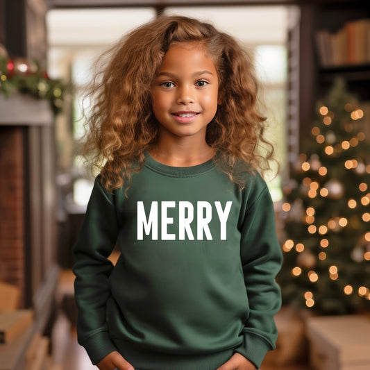 A young girl with curly hair smiles, wearing our MERRY • Kids Sweatshirt in green with white lettering. She stands indoors by a decorated Christmas tree and festive décor, showcasing this made-to-order holiday favorite.