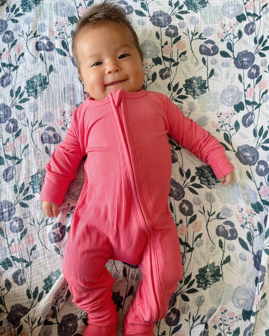 A smiling baby wears the Bright Pink Ribbed Solid • Bamboo Zippy (Updated Fit) onesie and lies on a floral-patterned blanket—perfect for sensitive skin.