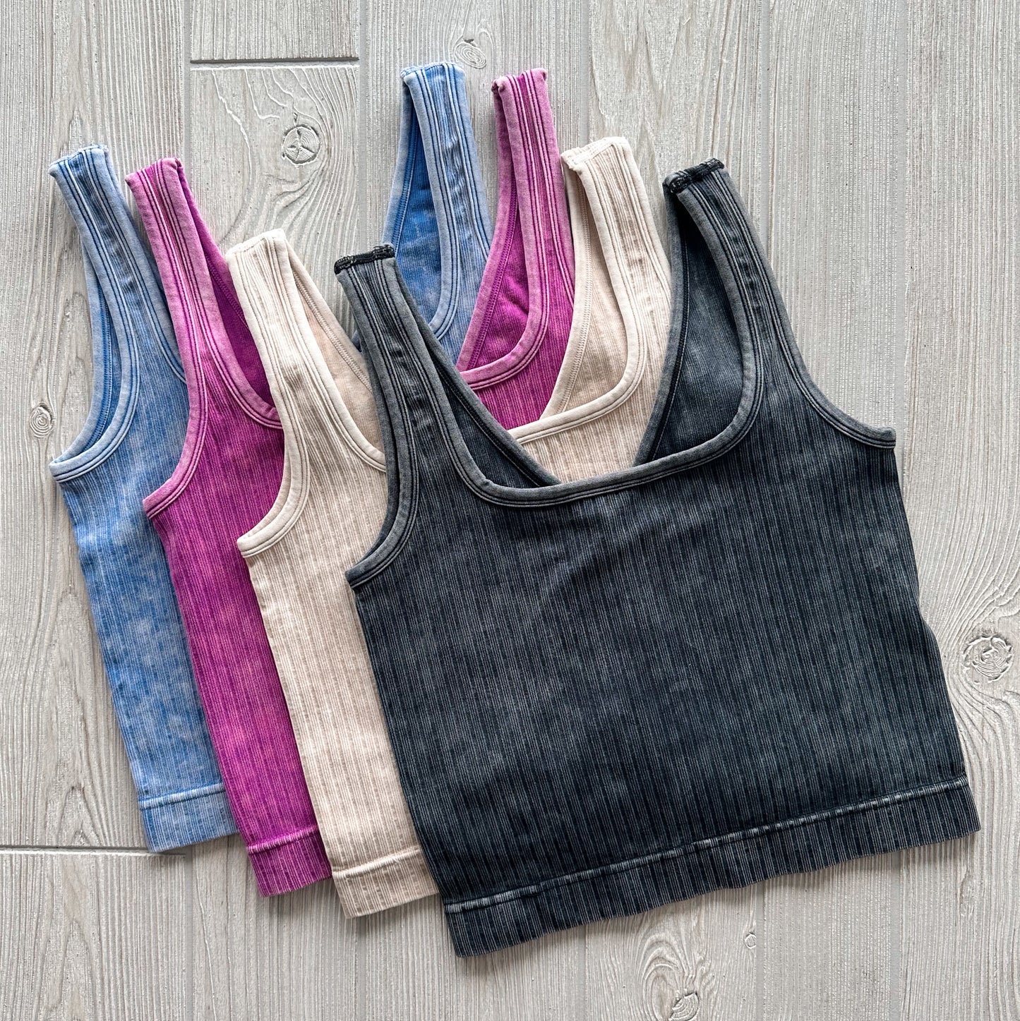 The Ava Ribbed Crop • Vintage Collection features five soft, stretchy brami tank tops in blue, magenta, beige, light beige, and black, neatly laid side by side on a light wooden surface.