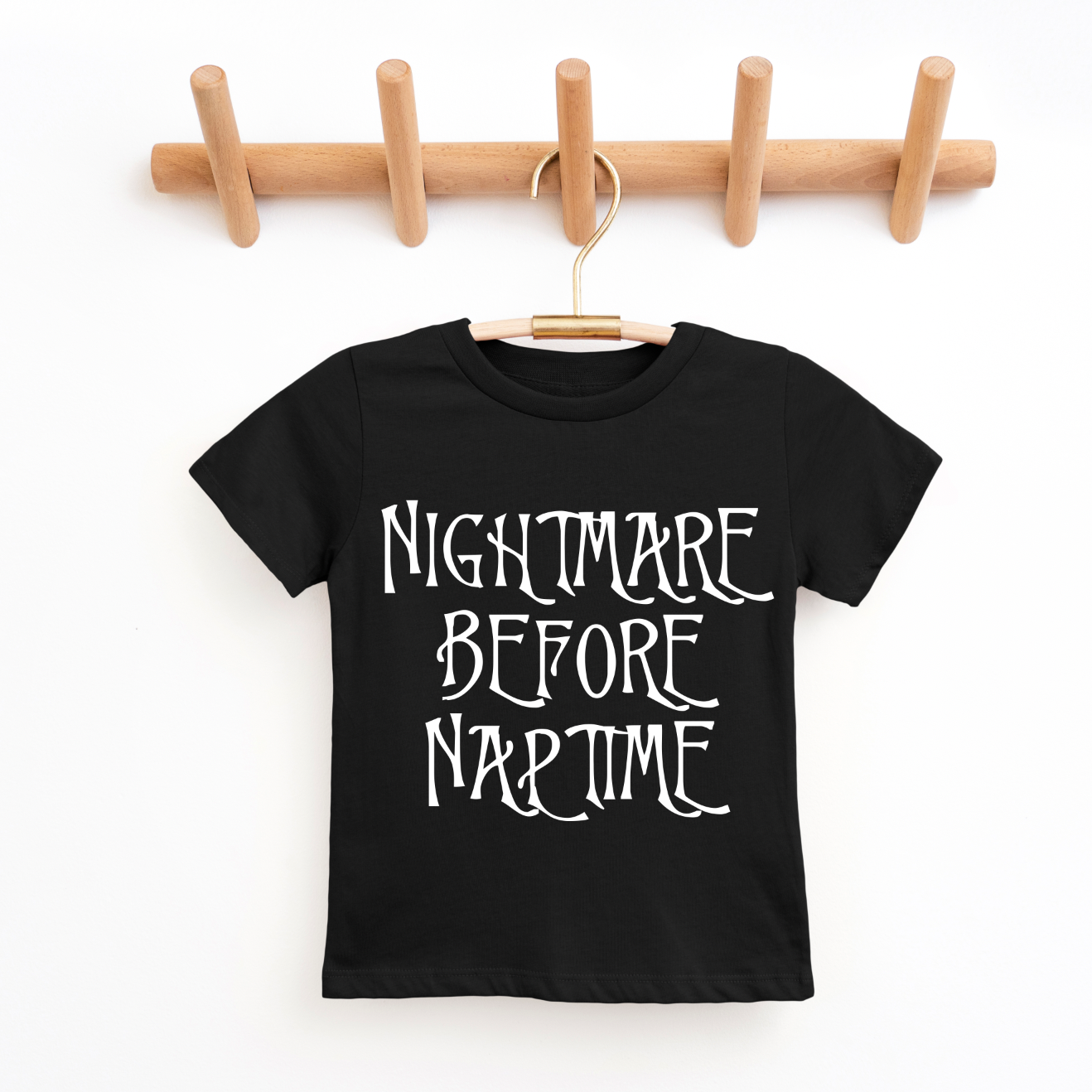 The Nightmare Before Naptime • Kids Tee is a black unisex shirt for kids, featuring “NIGHTMARE BEFORE NAPTIME” in white letters. It hangs on a wooden peg rack and offers a true-to-size fit—perfect for your little one! Shop small today.