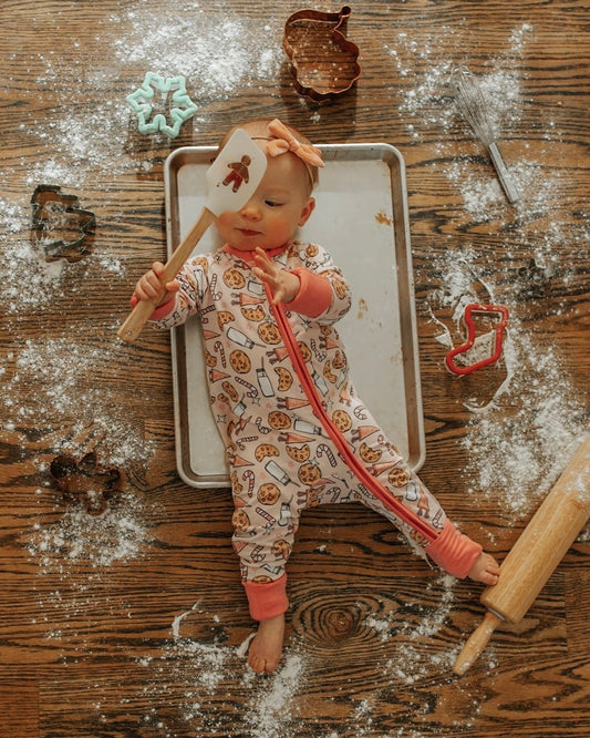A baby wearing the Milk & Cookies • Bamboo Zippy (Updated Fit) lies on a baking sheet atop a flour-dusted floor, holding a spatula. Cookie cutters, a rolling pin, and whisk surround them for a playful baking-inspired scene.