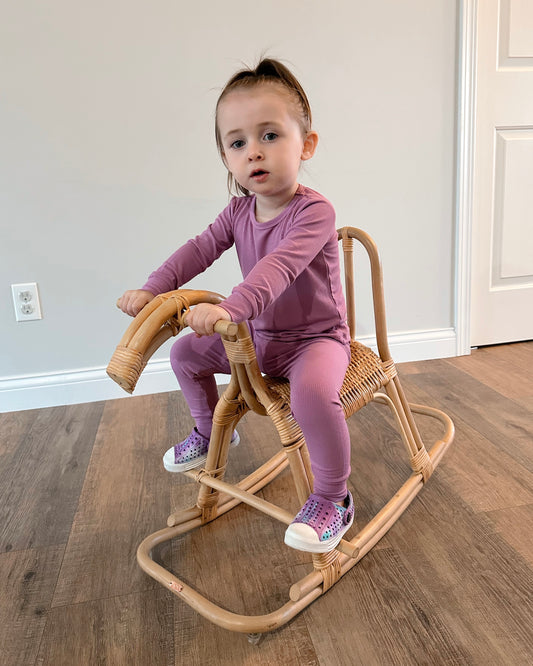 A young child with brown hair in a small ponytail wears the Lilac Ribbed Solid • 2 Piece Bamboo Set (Updated Fit) and colorful shoes while sitting on a wicker rocking horse indoors. The room has light walls and wood floors.