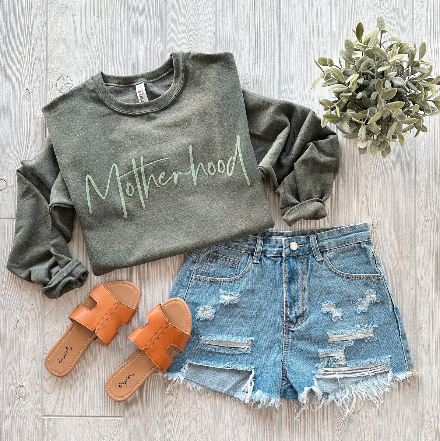 An olive Motherhood tonal pullover, distressed denim shorts, tan slide sandals, and a small green potted plant styled on a light wood floor.