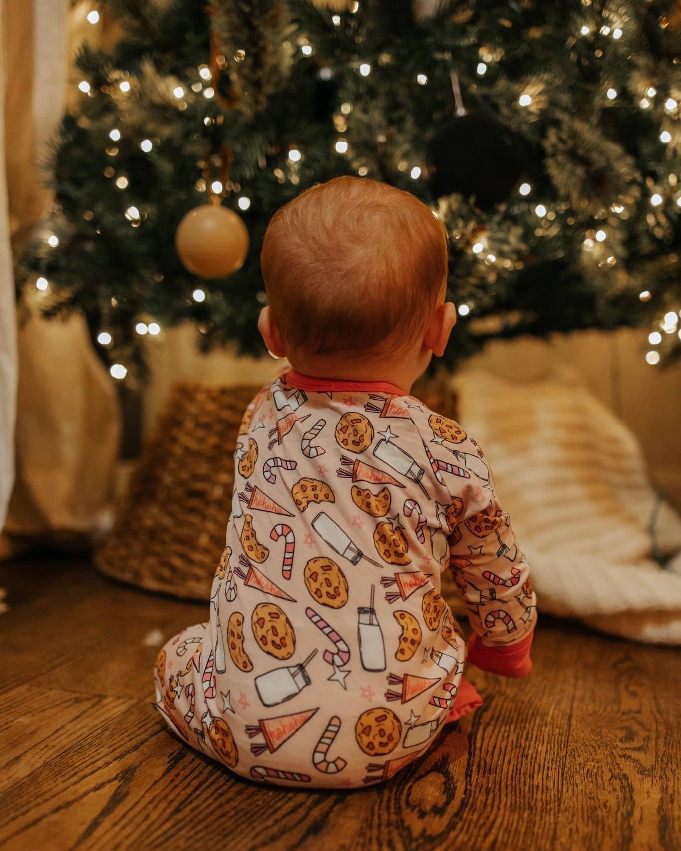A baby in the Milk & Cookies • Bamboo Zippy (Updated Fit), featuring soft bamboo viscose and festive cookie, milk, and candy cane designs, sits on a wooden floor facing a decorated Christmas tree.