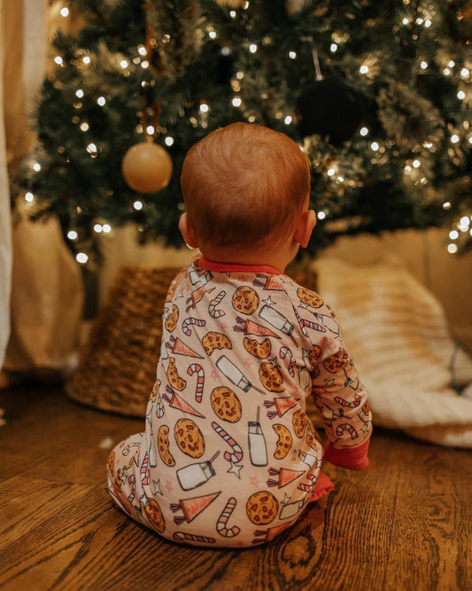 A baby in the Milk & Cookies • Bamboo Zippy (Updated Fit), featuring soft bamboo viscose and festive cookie, milk, and candy cane designs, sits on a wooden floor facing a decorated Christmas tree.