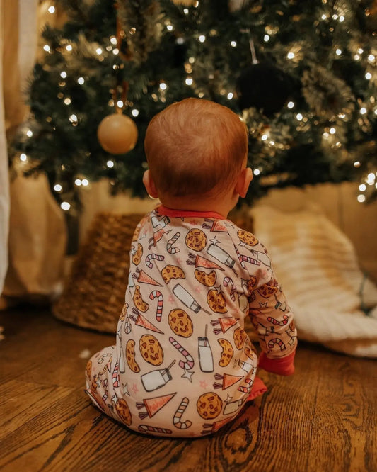 A baby in the Milk & Cookies • Bamboo Zippy (Updated Fit), featuring soft bamboo viscose and festive cookie, milk, and candy cane designs, sits on a wooden floor facing a decorated Christmas tree.
