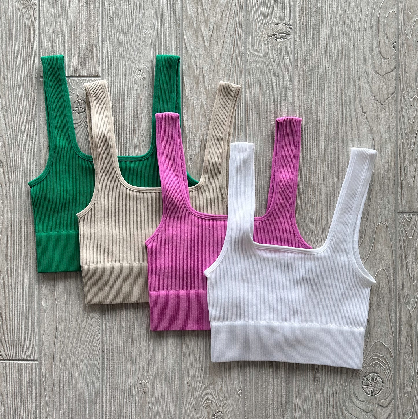 Four Iris Crop tops in green, beige, pink, and white are displayed on a light wooden floor. Made from stretchy fabric with wide straps and a square neckline, theyre ideal for layering or wearing as comfy lounge bras.