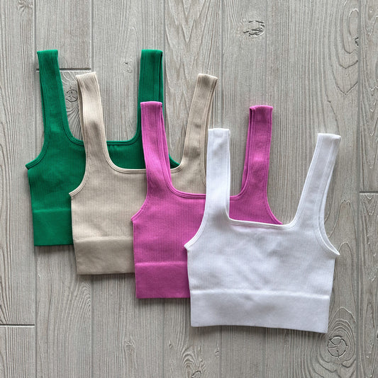 Four Iris Crop tops in green, beige, pink, and white are displayed on a light wooden floor. Made from stretchy fabric with wide straps and a square neckline, theyre ideal for layering or wearing as comfy lounge bras.