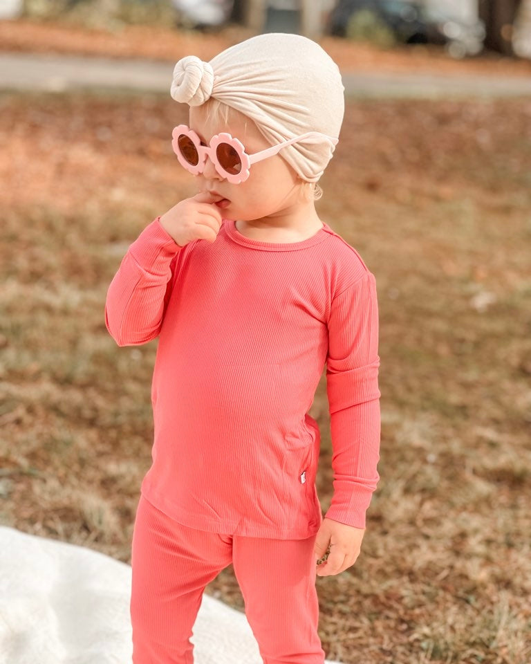 A toddler wears the Bright Pink Ribbed Solid • 2 Piece Bamboo Set (Updated Fit) with flower sunglasses and a cream head wrap, standing on grass against a softly blurred brown and green outdoor background.